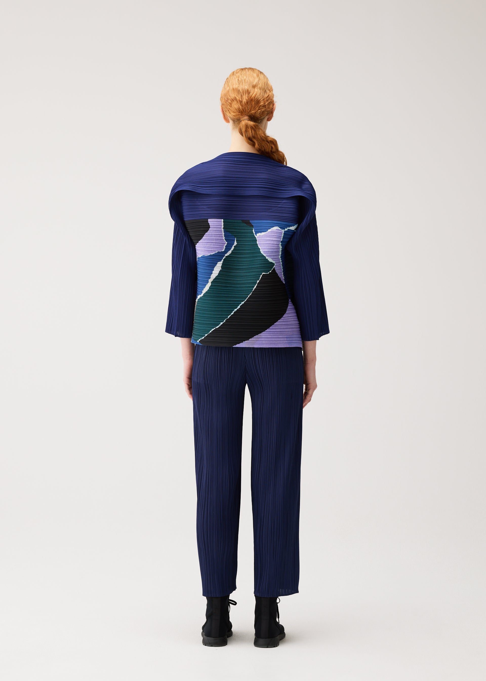 Zoomed view A person with red hair in a low ponytail wears PLEATS PLEASE ISSEY MIYAKE’s SURPRISE! — a blue pleated outfit featuring a unique design with a colorful abstract pattern on the back. They are standing against a plain light background, facing away from the camera.