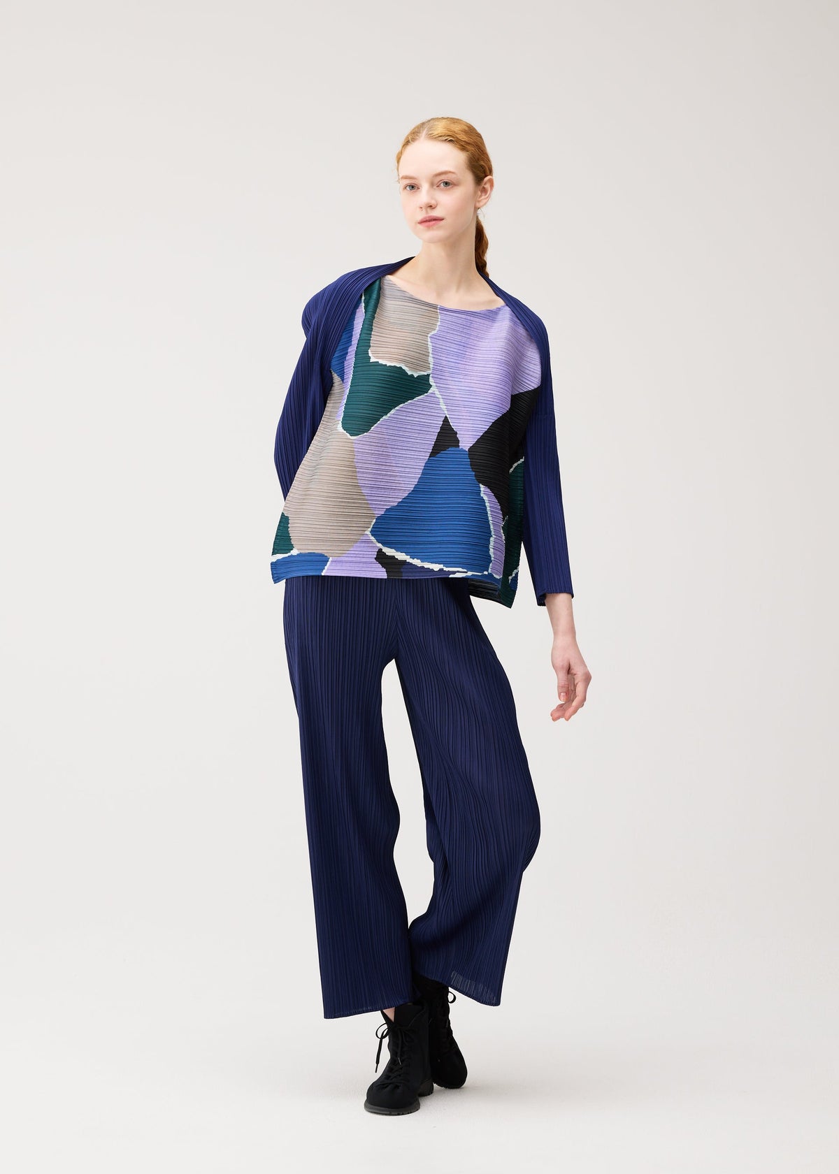 A person stands in a relaxed pose, wearing the SURPRISE! pleated outfit by PLEATS PLEASE ISSEY MIYAKE, featuring a loose boat-neck top adorned with abstract blue, purple, and green shapes and matching navy wide-leg pants. The background is plain and light-colored. view 7 of 7