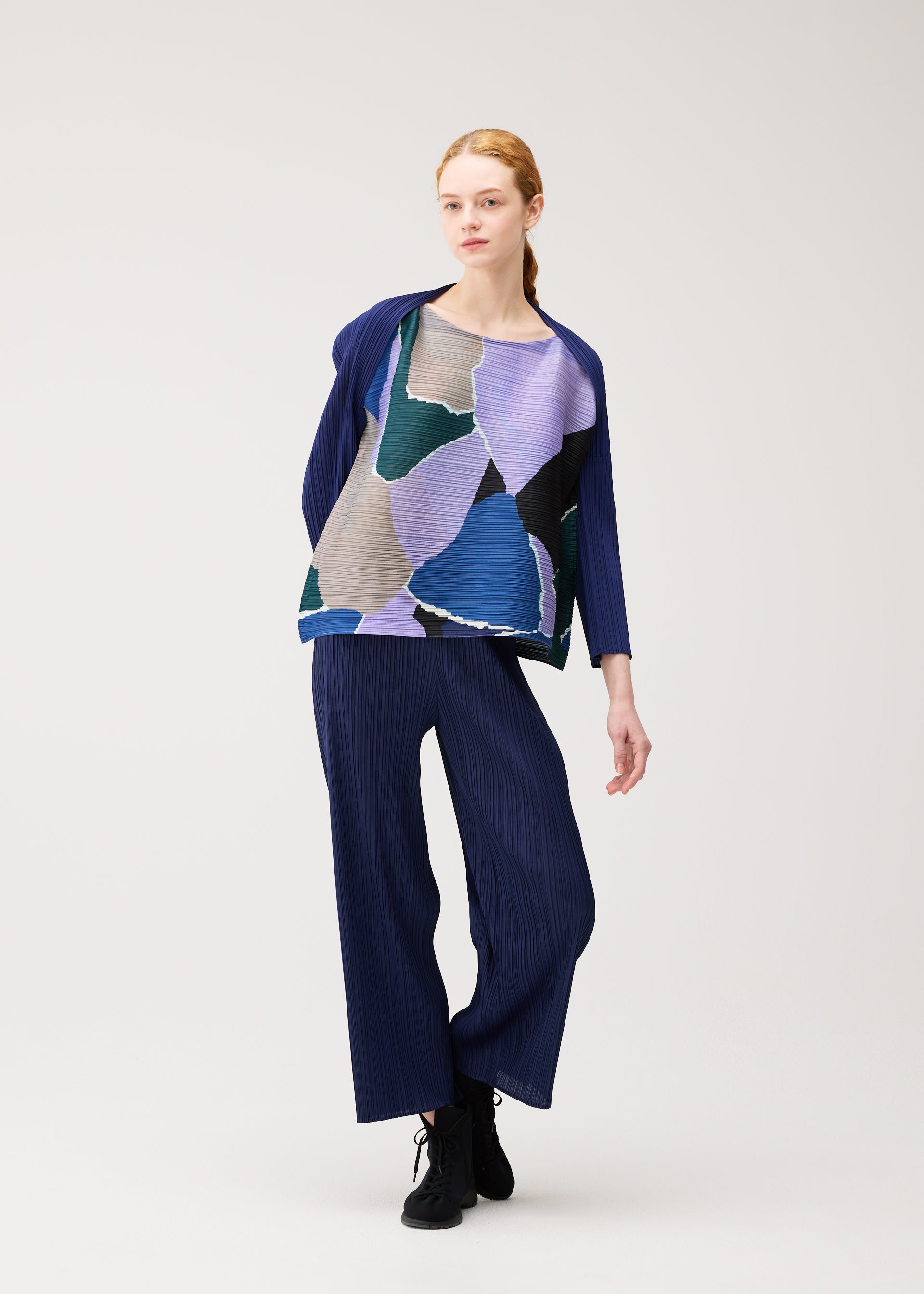 Zoomed view A person stands in a relaxed pose, wearing the SURPRISE! pleated outfit by PLEATS PLEASE ISSEY MIYAKE, featuring a loose boat-neck top adorned with abstract blue, purple, and green shapes and matching navy wide-leg pants. The background is plain and light-colored.