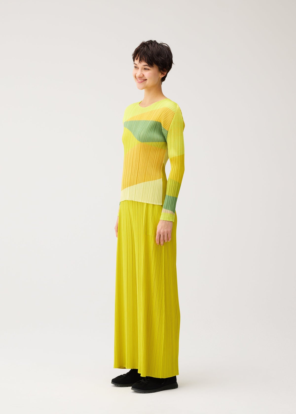 A person models the ORBIT set by PLEATS PLEASE ISSEY MIYAKE—featuring a long, pleated yellow skirt and a matching yellow-green, round-neck long-sleeved top—against a plain white background. view 5 of 7