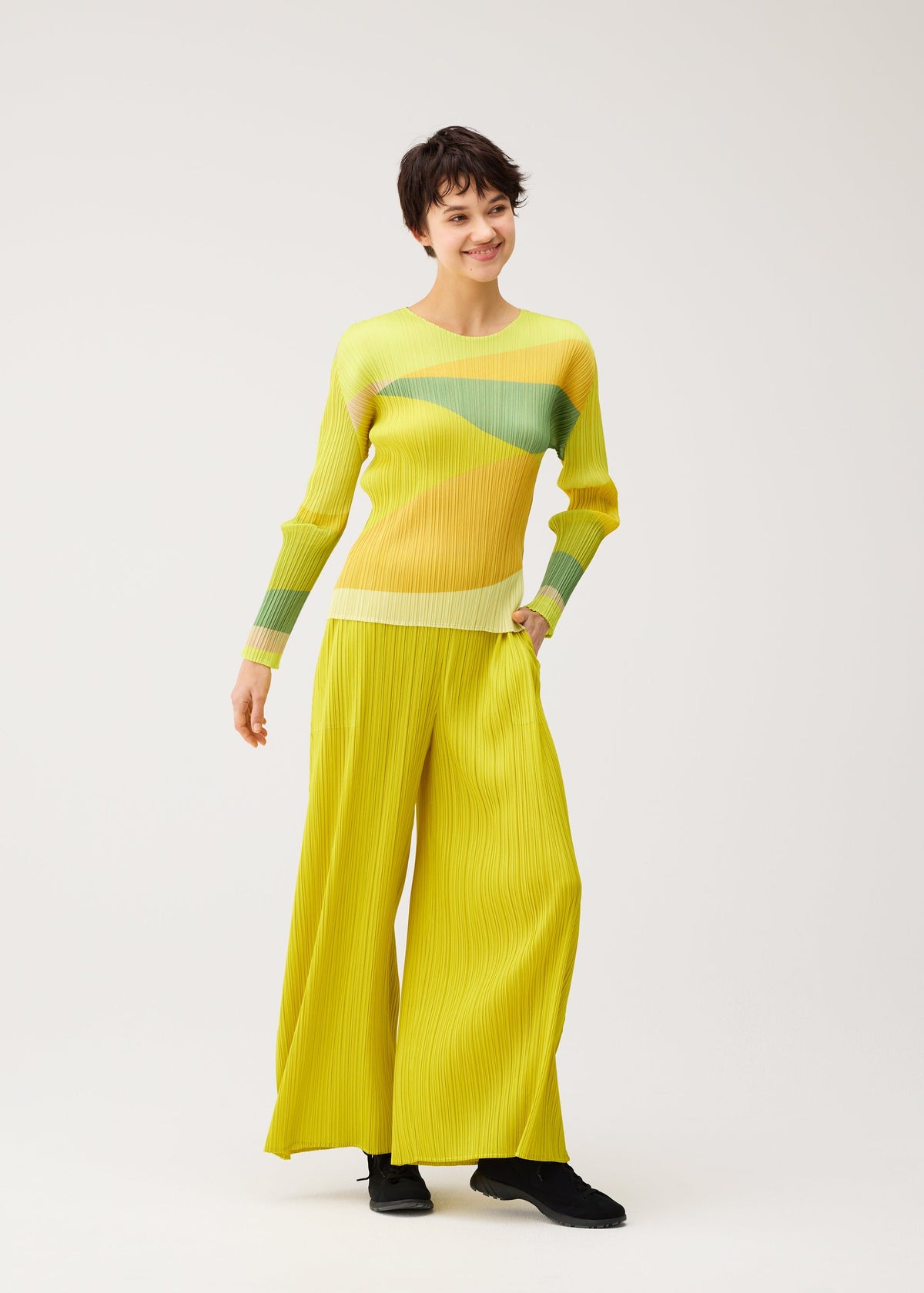 A person smiles in the ORBIT top by PLEATS PLEASE ISSEY MIYAKE, featuring sheer fabric with yellow, green, and beige stripes, paired with bright yellow wide-leg pants and black shoes against a white backdrop. view 7 of 7