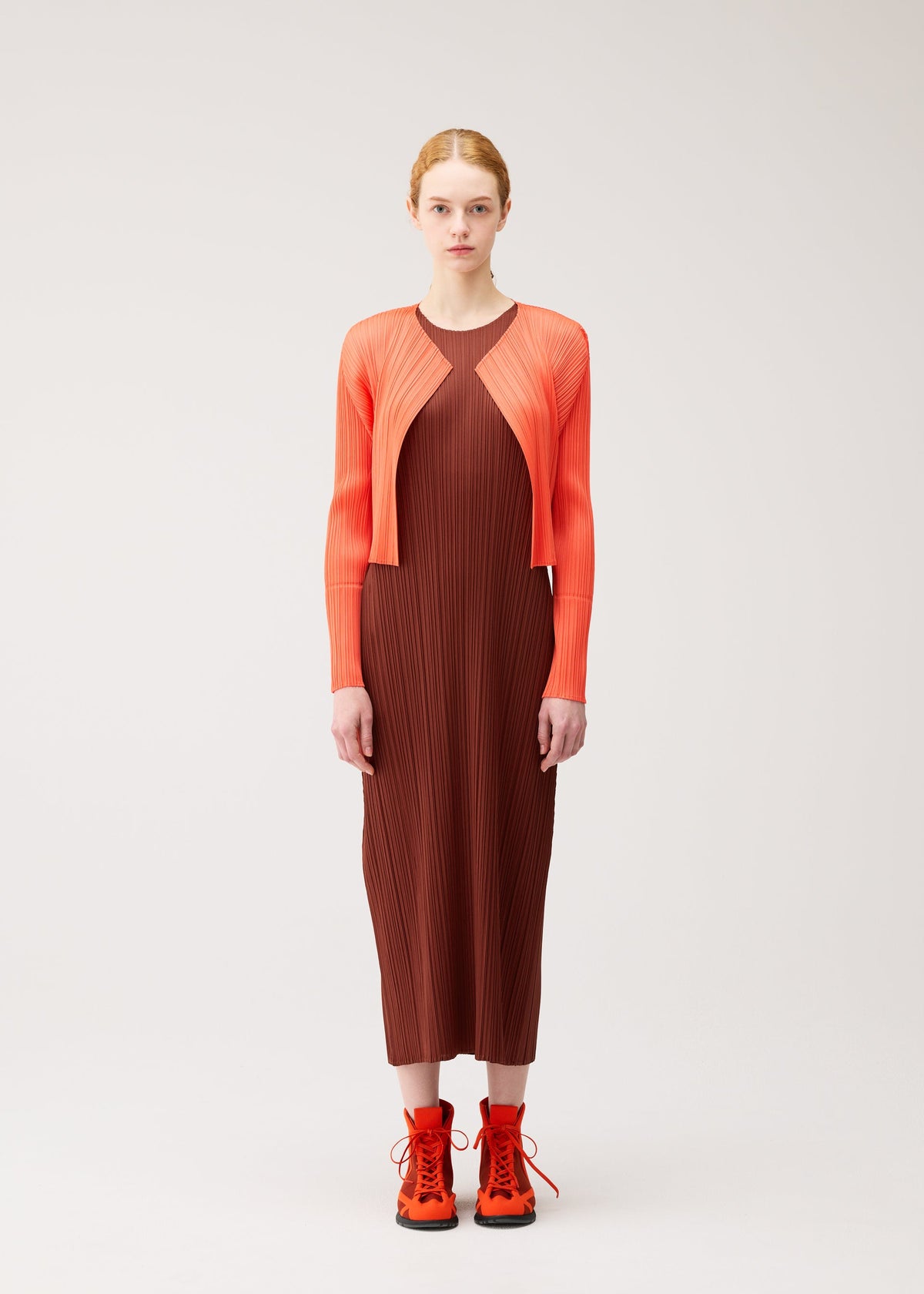 A person stands facing forward against a plain, light background, effortlessly showcasing gentle tones and seasonal colors dressed in the MONTHLY COLORS : SEPTEMBER collection by PLEATS PLEASE ISSEY MIYAKE, featuring a brown pleated dress paired with an orange cardigan and orange lace-up shoes. view 7 of 10