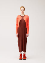 A person stands facing forward against a plain, light background, effortlessly showcasing gentle tones and seasonal colors dressed in the MONTHLY COLORS : SEPTEMBER collection by PLEATS PLEASE ISSEY MIYAKE, featuring a brown pleated dress paired with an orange cardigan and orange lace-up shoes.