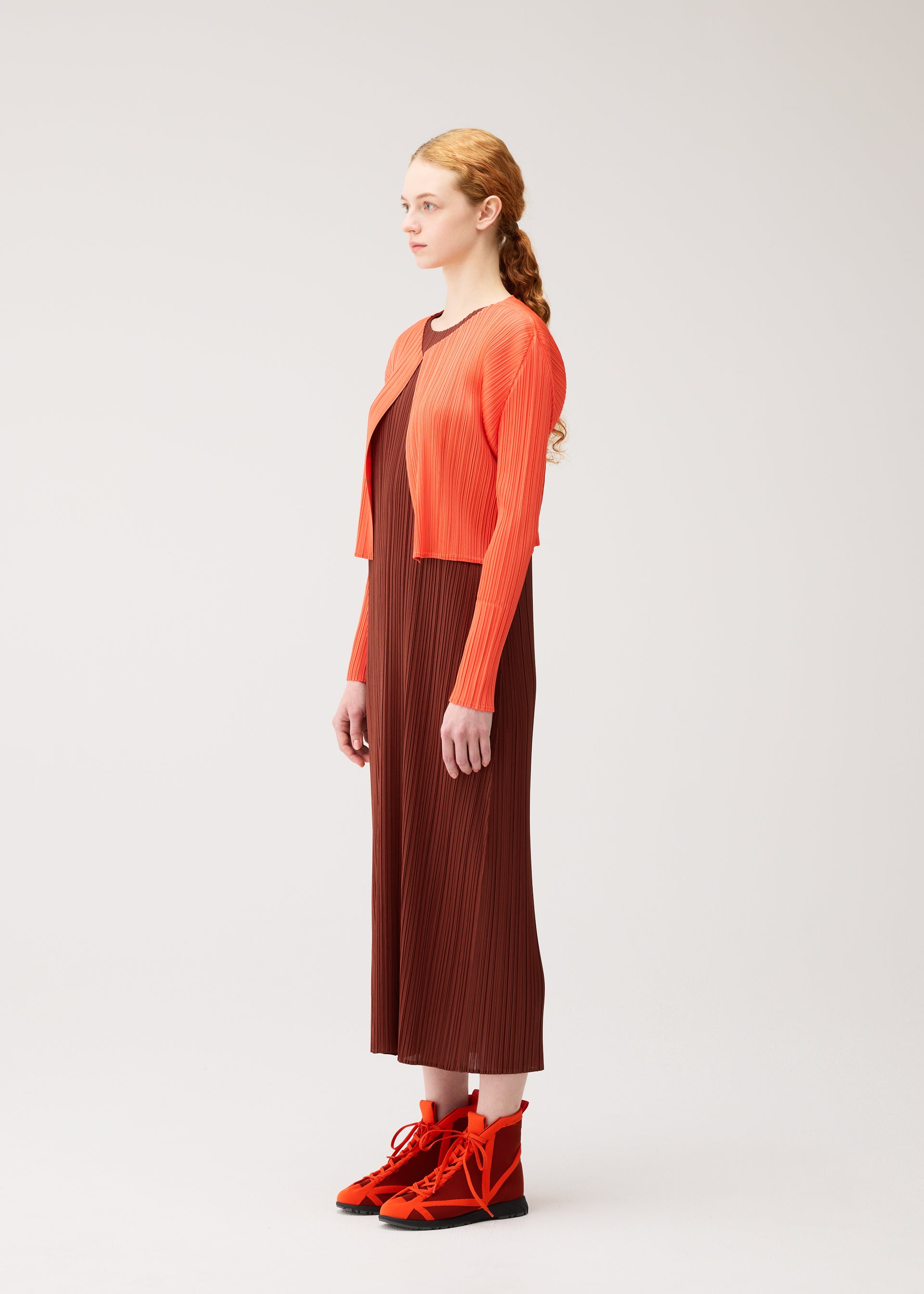 Zoomed view A woman with long hair in a low ponytail stands sideways against a plain light background, wearing the PLEATS PLEASE ISSEY MIYAKE MONTHLY COLORS : SEPTEMBER brown pleated dress styled with an orange cropped cardigan and orange lace-up boots in gentle tones.