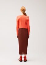 A person with long, reddish hair styled in a low ponytail wears the MONTHLY COLORS : SEPTEMBER collection from PLEATS PLEASE ISSEY MIYAKE, featuring a textured orange top beneath a soft cardigan, paired with a brown pleated skirt and orange lace-up shoes. The look embraces gentle tones and subtle seasonal colors as they stand facing away against a plain background.