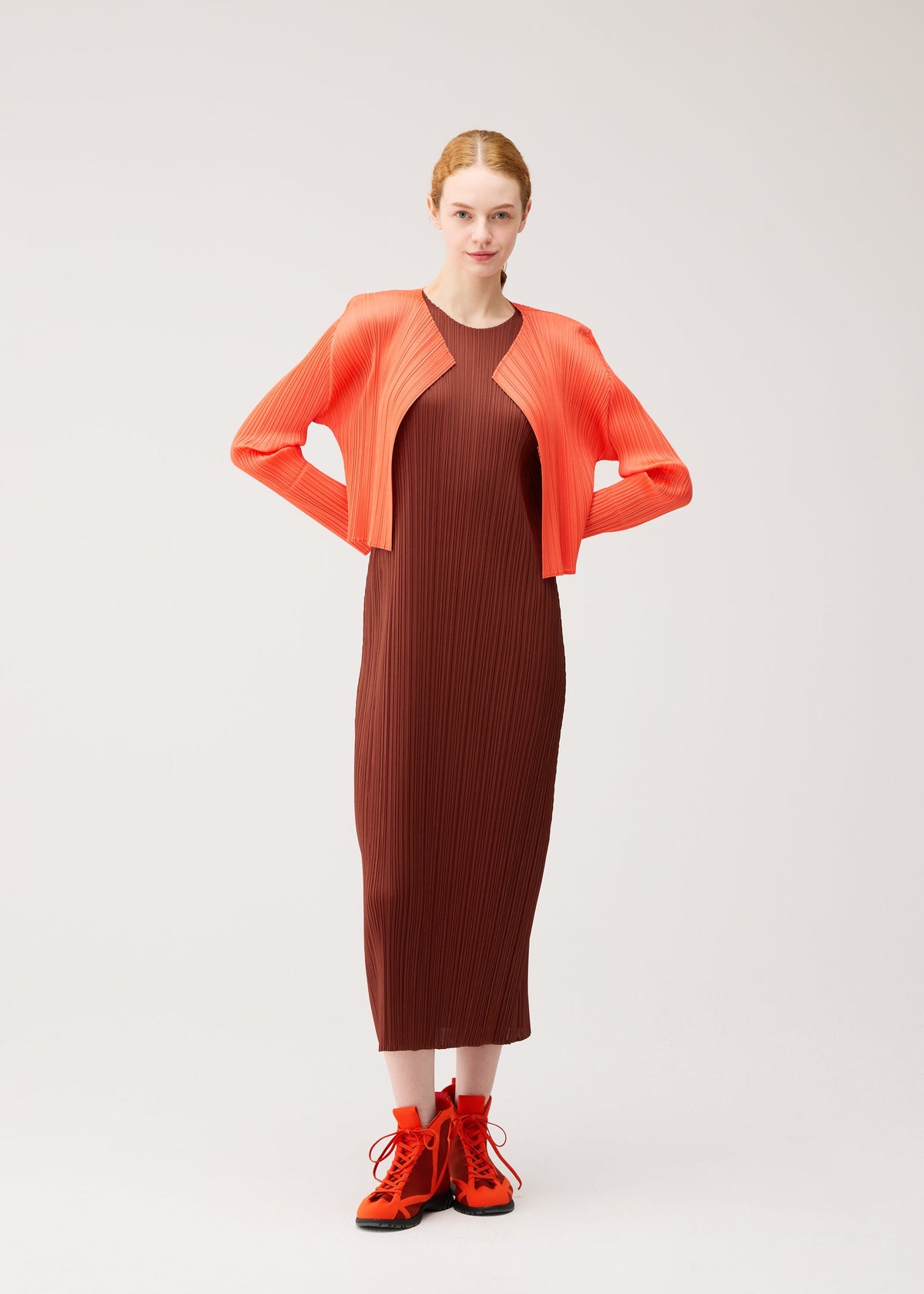 A woman stands against a plain background wearing an ankle-length, pleated dress in soft brown tones from the PLEATS PLEASE ISSEY MIYAKE MONTHLY COLORS : SEPTEMBER collection, styled with a vibrant orange cropped jacket and matching boots. Her hair is tied back and her hands rest on her hips. view 10 of 10