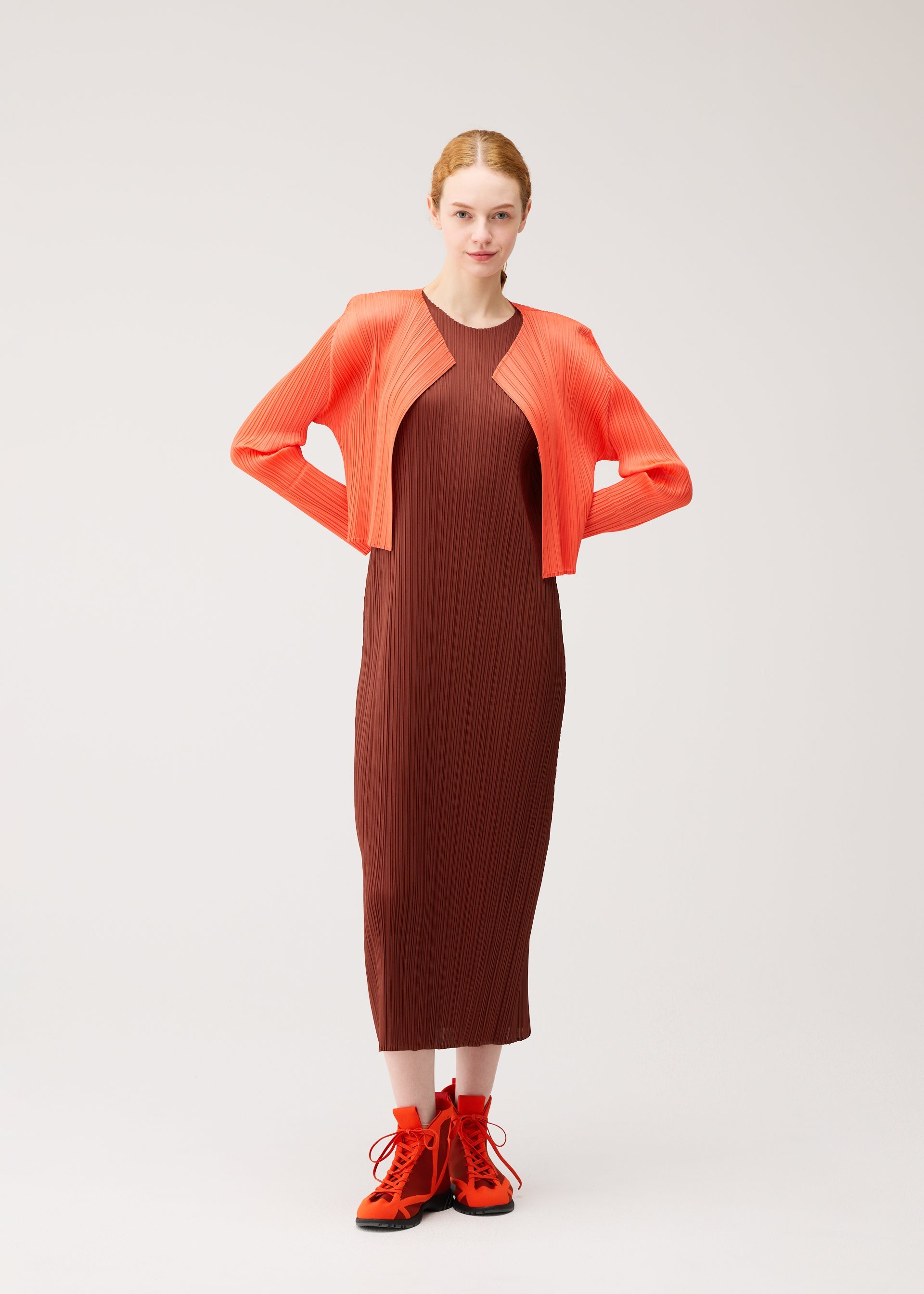Zoomed view A woman stands against a plain background wearing an ankle-length, pleated dress in soft brown tones from the PLEATS PLEASE ISSEY MIYAKE MONTHLY COLORS : SEPTEMBER collection, styled with a vibrant orange cropped jacket and matching boots. Her hair is tied back and her hands rest on her hips.
