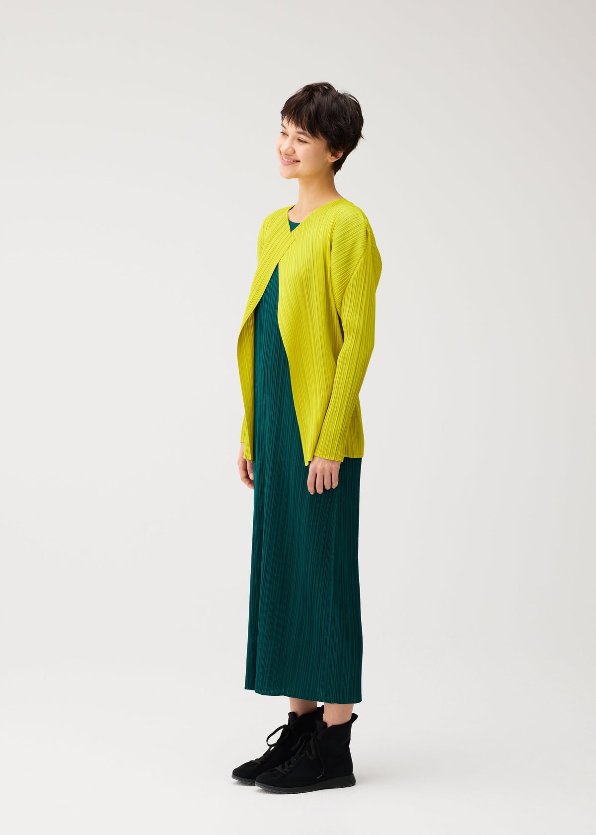 Zoomed view A person with short dark hair smiles in the MONTHLY COLORS : OCTOBER dress by PLEATS PLEASE ISSEY MIYAKE, styled with a yellow color block cardigan and black ankle boots against a white background.