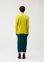 A person with short dark hair faces away, wearing the MONTHLY COLORS : OCTOBER by PLEATS PLEASE ISSEY MIYAKE—a bright yellow pleated top under a dark green pleated skirt with black ankle boots against a plain background.
