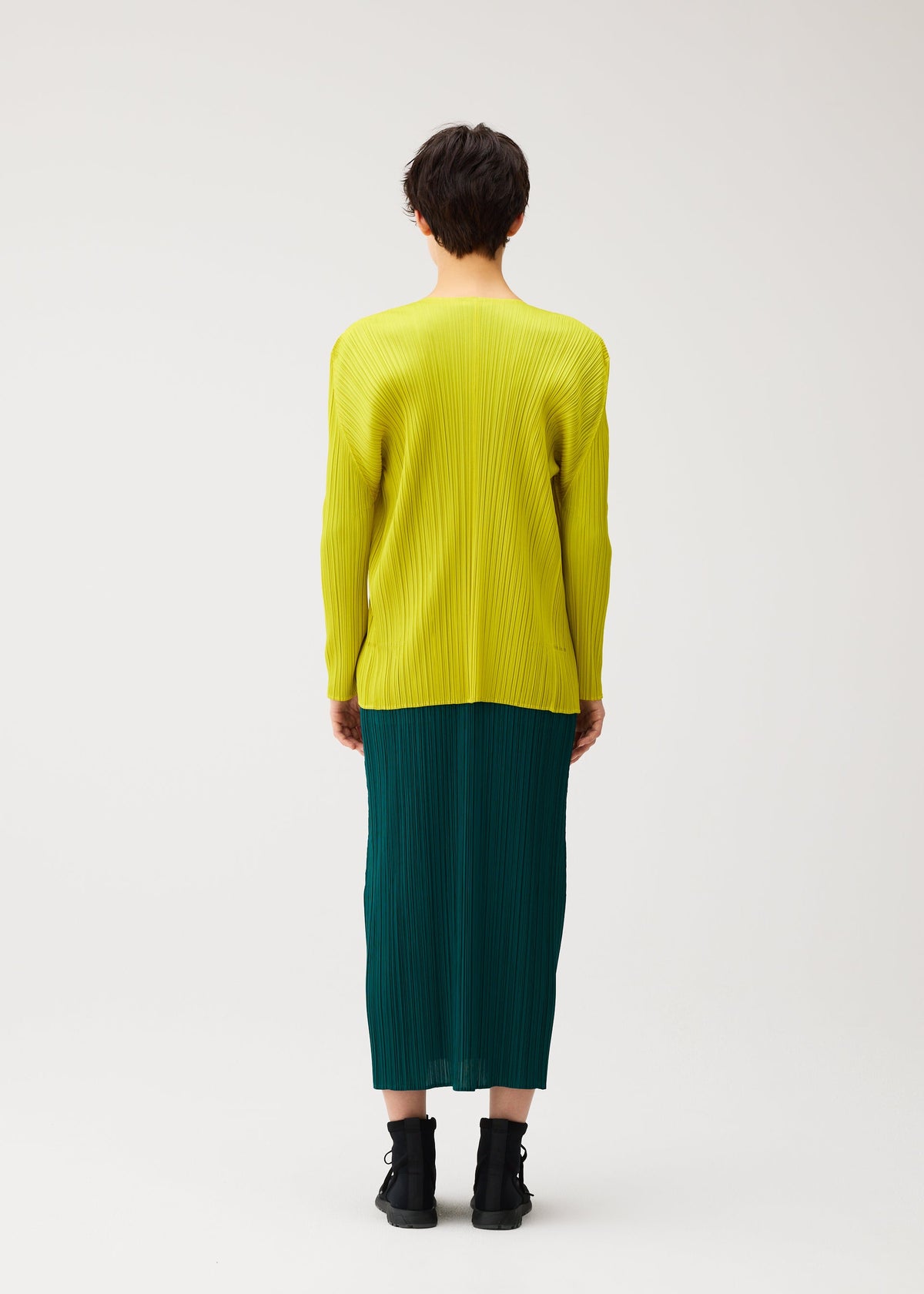 Zoomed view A person with short dark hair faces away, wearing the MONTHLY COLORS : OCTOBER by PLEATS PLEASE ISSEY MIYAKE—a bright yellow pleated top under a dark green pleated skirt with black ankle boots against a plain background.