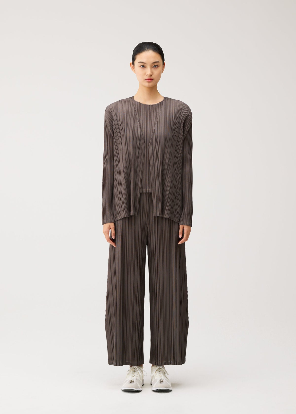 A person stands facing forward, dressed in the MELLOW PLEATS set by PLEATS PLEASE ISSEY MIYAKE—a loose, pleated brown top and matching wide-leg pants with handy pockets—paired with white sneakers against a plain light background. view 6 of 9