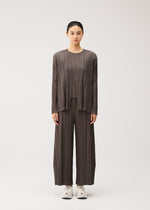 A person stands facing forward, dressed in the MELLOW PLEATS set by PLEATS PLEASE ISSEY MIYAKE—a loose, pleated brown top and matching wide-leg pants with handy pockets—paired with white sneakers against a plain light background.