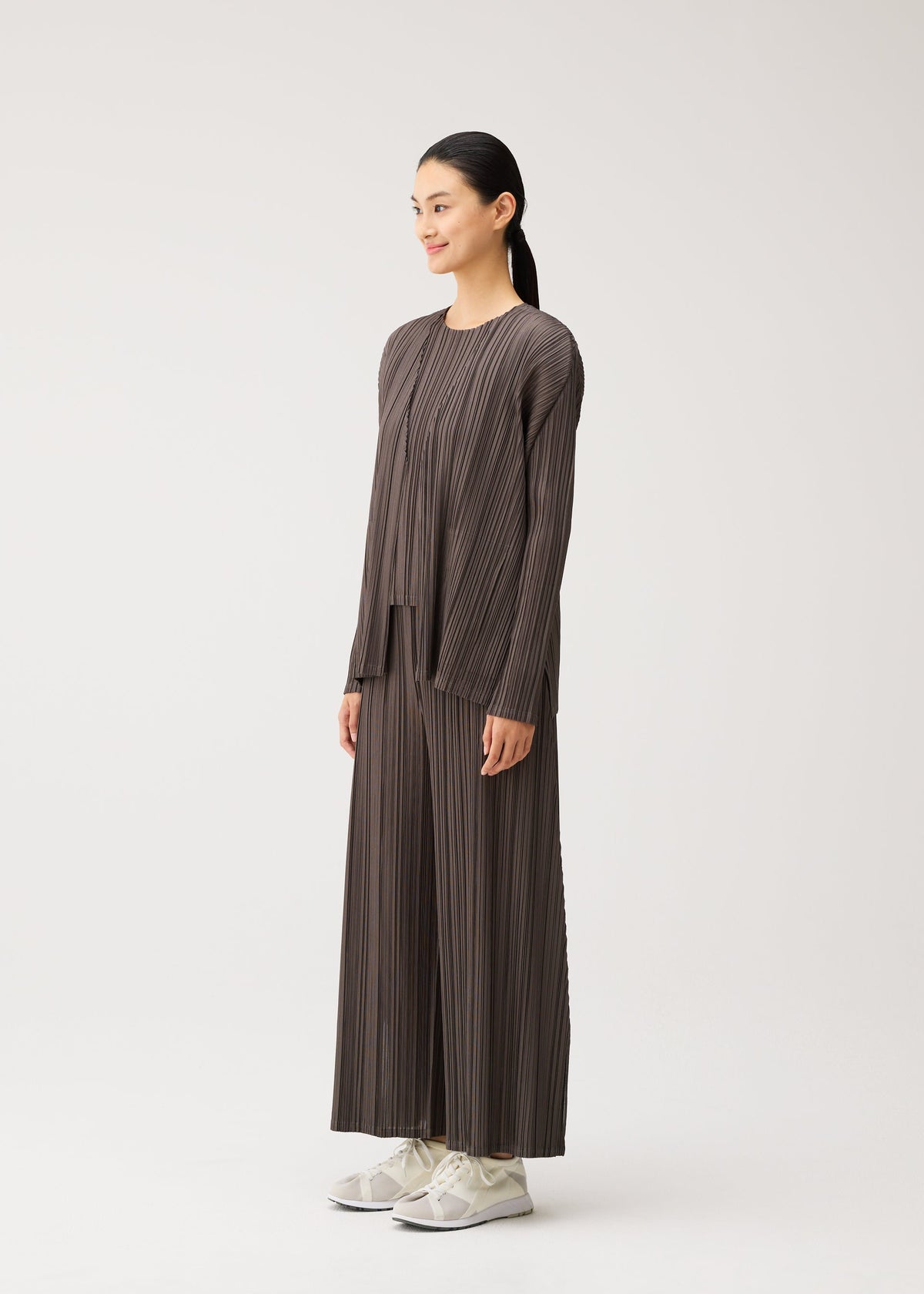 A woman stands sideways against a plain white background, wearing the MELLOW PLEATS matching set by PLEATS PLEASE ISSEY MIYAKE—featuring a loose-fitting, pleated brown top and wide-leg pants with pockets—paired with white sneakers. view 7 of 9