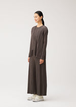 A woman stands sideways against a plain white background, wearing the MELLOW PLEATS matching set by PLEATS PLEASE ISSEY MIYAKE—featuring a loose-fitting, pleated brown top and wide-leg pants with pockets—paired with white sneakers.