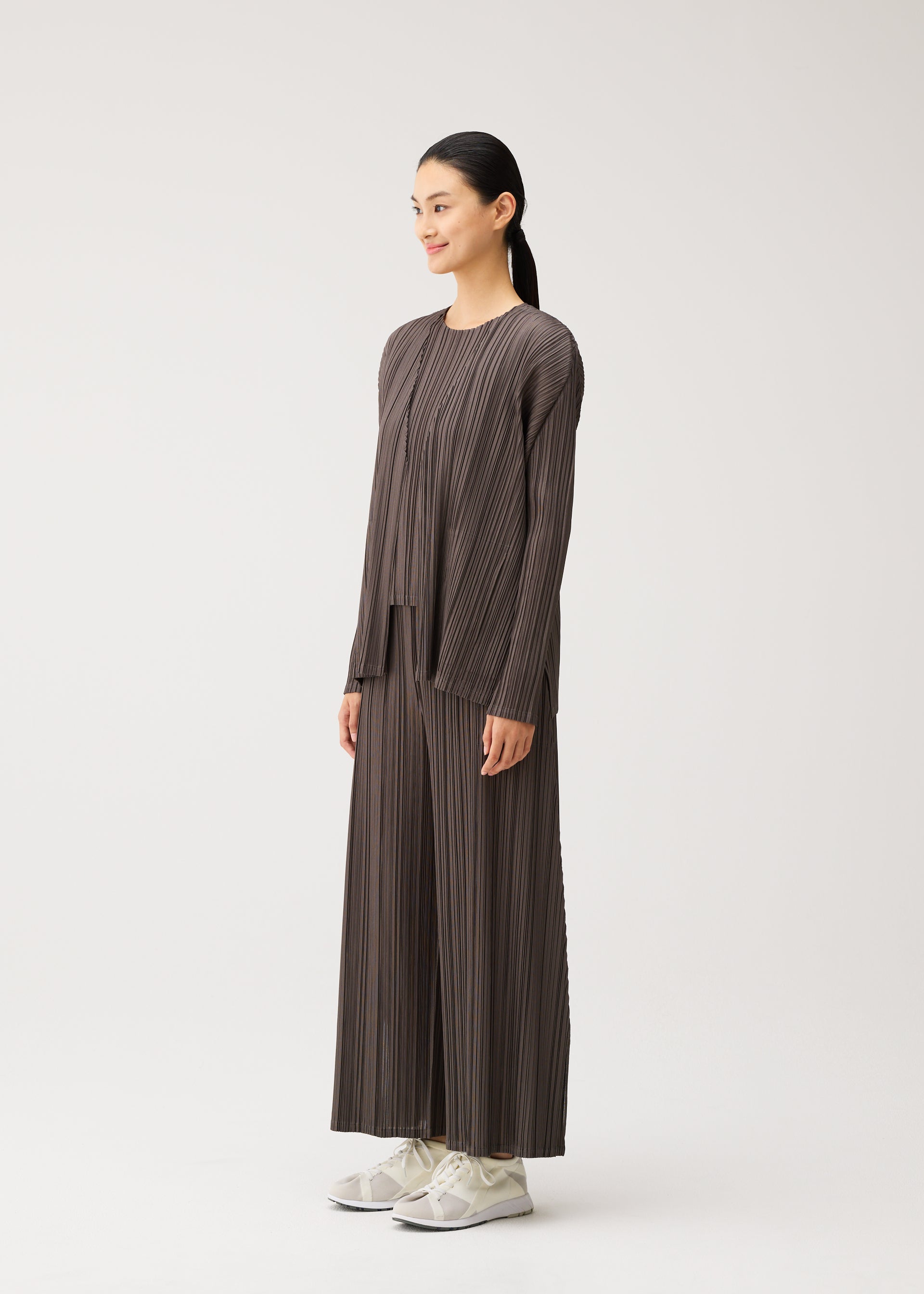 Zoomed view A woman stands sideways against a plain white background, wearing the MELLOW PLEATS matching set by PLEATS PLEASE ISSEY MIYAKE—featuring a loose-fitting, pleated brown top and wide-leg pants with pockets—paired with white sneakers.