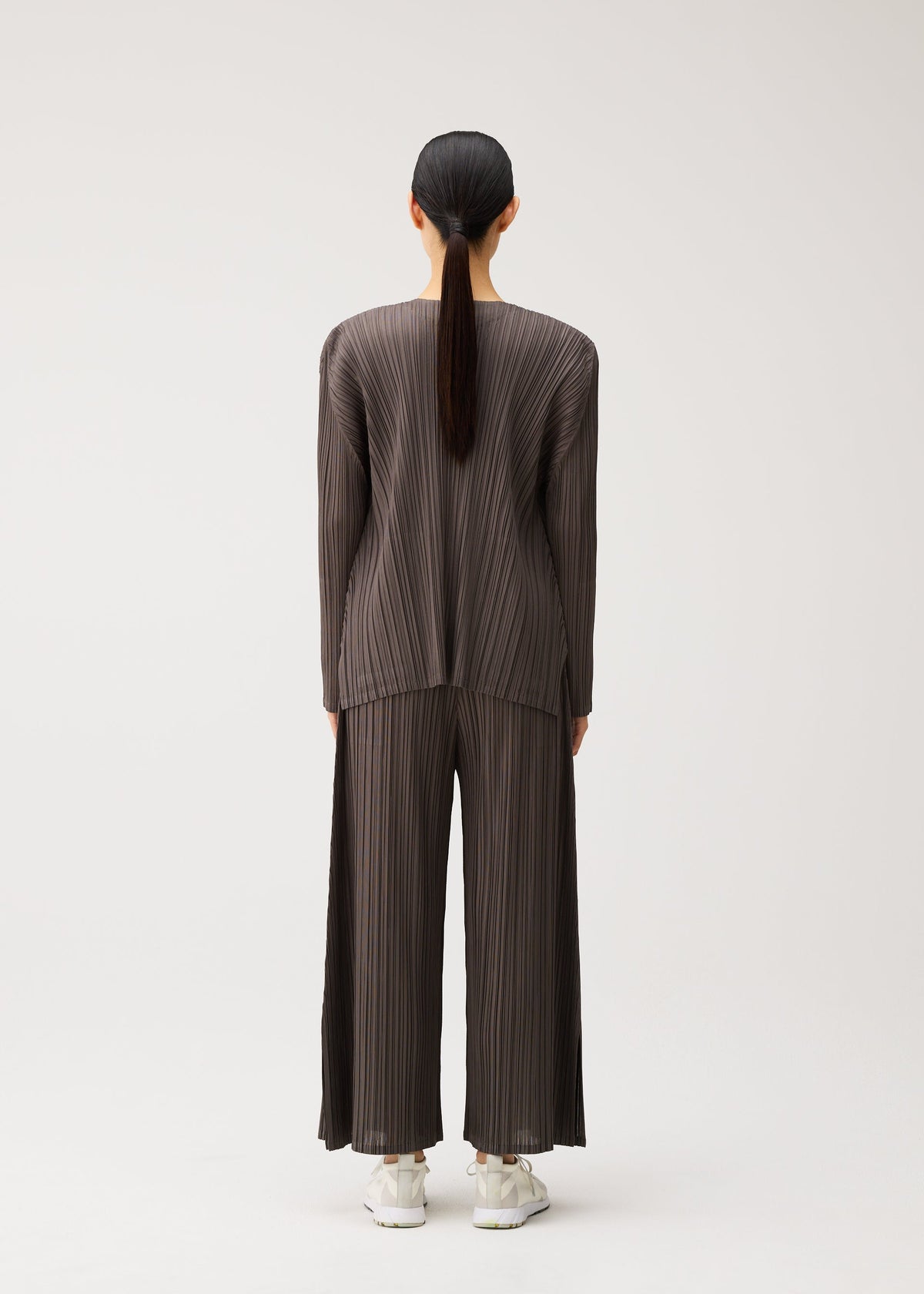 A person with long dark hair, seen from behind, wears the MELLOW PLEATS matching pleated brown top and wide-leg pants by PLEATS PLEASE ISSEY MIYAKE, crafted from a soft-textured fabric with pockets and paired with white sneakers, standing against a plain light background. view 8 of 9