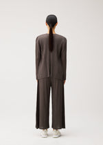 A person with long dark hair, seen from behind, wears the MELLOW PLEATS matching pleated brown top and wide-leg pants by PLEATS PLEASE ISSEY MIYAKE, crafted from a soft-textured fabric with pockets and paired with white sneakers, standing against a plain light background.