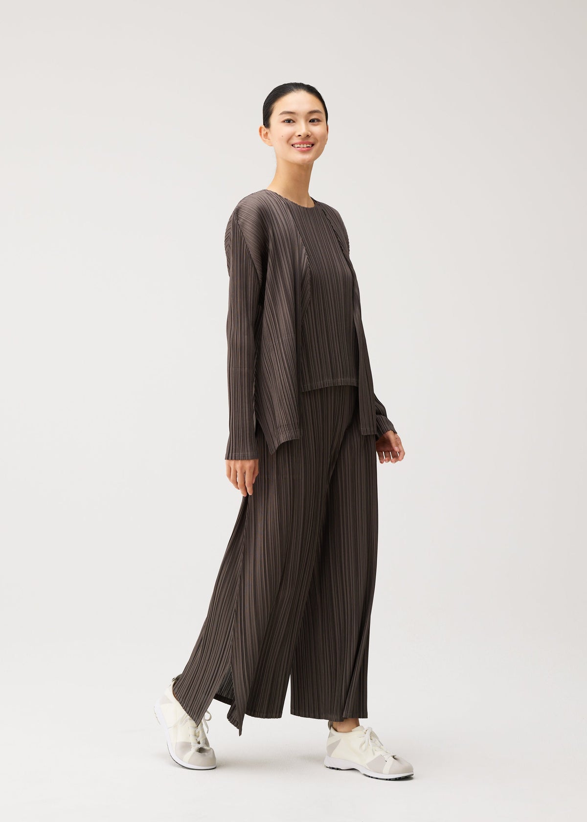 A person smiles while walking, dressed in the MELLOW PLEATS loose-fitting, dark pleated top and matching wide-leg pants with pockets from PLEATS PLEASE ISSEY MIYAKE, paired with white sneakers against a plain light background. view 9 of 9