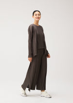 A person smiles while walking, dressed in the MELLOW PLEATS loose-fitting, dark pleated top and matching wide-leg pants with pockets from PLEATS PLEASE ISSEY MIYAKE, paired with white sneakers against a plain light background.