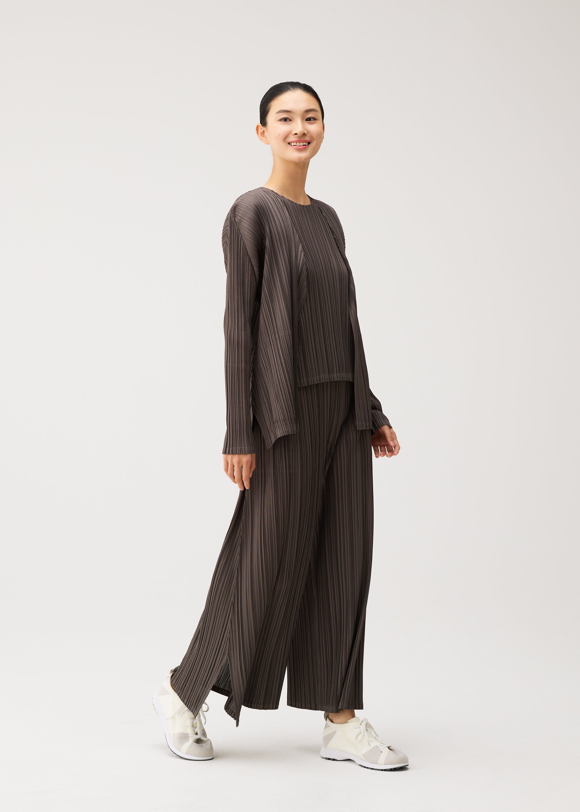 Zoomed view A person smiles while walking, dressed in the MELLOW PLEATS loose-fitting, dark pleated top and matching wide-leg pants with pockets from PLEATS PLEASE ISSEY MIYAKE, paired with white sneakers against a plain light background.