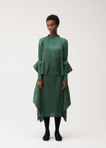 A woman stands facing forward against a plain light background, wearing the FLUTTER dress by PLEATS PLEASE ISSEY MIYAKE—a green, pleated, long-sleeved piece featuring an asymmetrical hem—styled with a distinctive cardigan draped over her shoulders. She pairs the look with black tights and black shoes, maintaining a neutral expression.