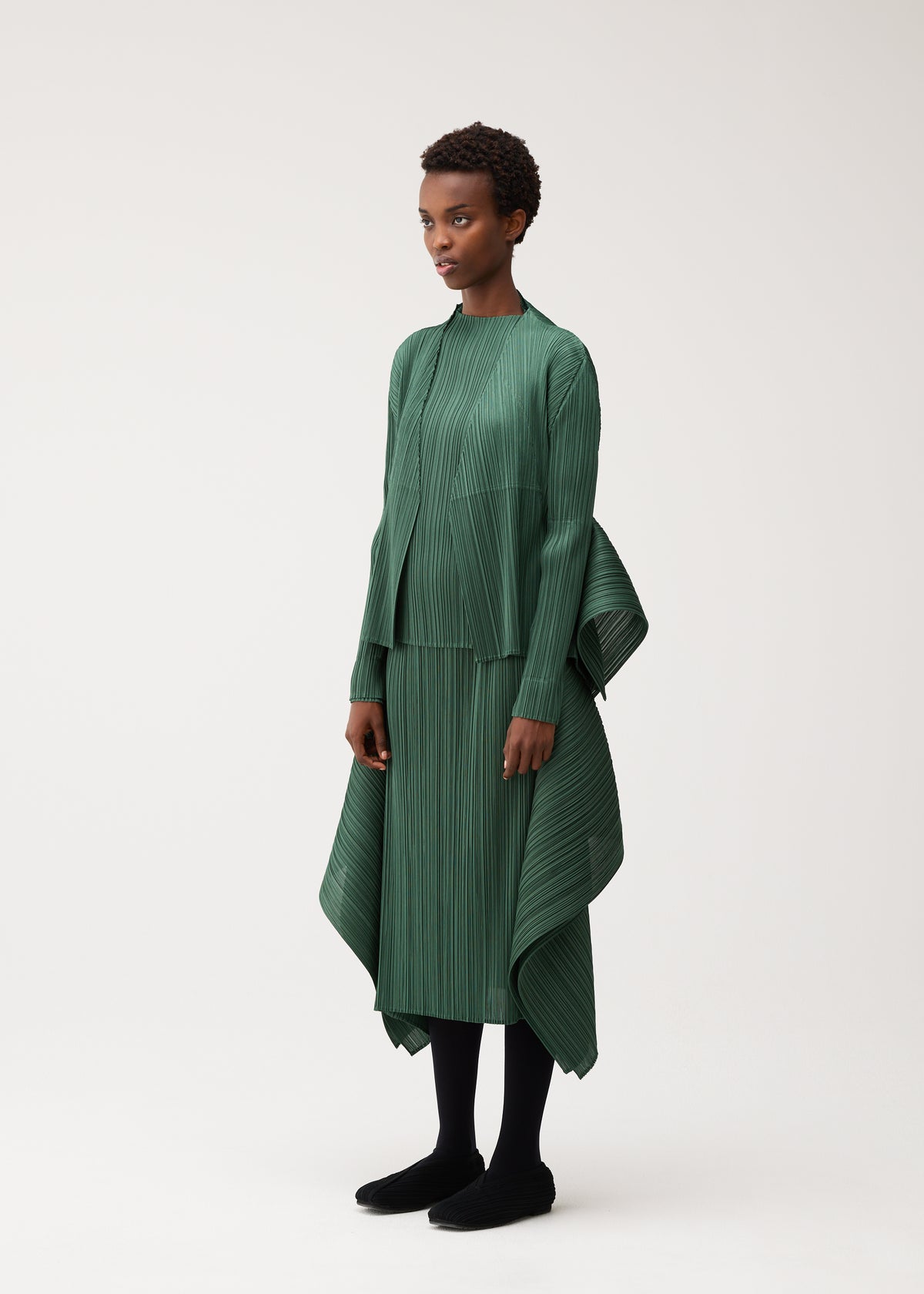A person stands wearing FLUTTER, a green pleated asymmetrical dress with long sleeves and a high-low hem from PLEATS PLEASE ISSEY MIYAKE, styled with a unique long-sleeved cardigan. The look is completed with black leggings and black shoes against a plain light background. view 7 of 9