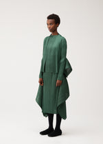 A person stands wearing FLUTTER, a green pleated asymmetrical dress with long sleeves and a high-low hem from PLEATS PLEASE ISSEY MIYAKE, styled with a unique long-sleeved cardigan. The look is completed with black leggings and black shoes against a plain light background.