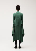 A person stands with their back to the camera, wearing the FLUTTER dress by PLEATS PLEASE ISSEY MIYAKE—a long, pleated dark green piece featuring draped, layered fabric. The outfit is paired with a distinctive cardigan for added texture and interest, along with black tights and shoes. The background is plain and light-colored.