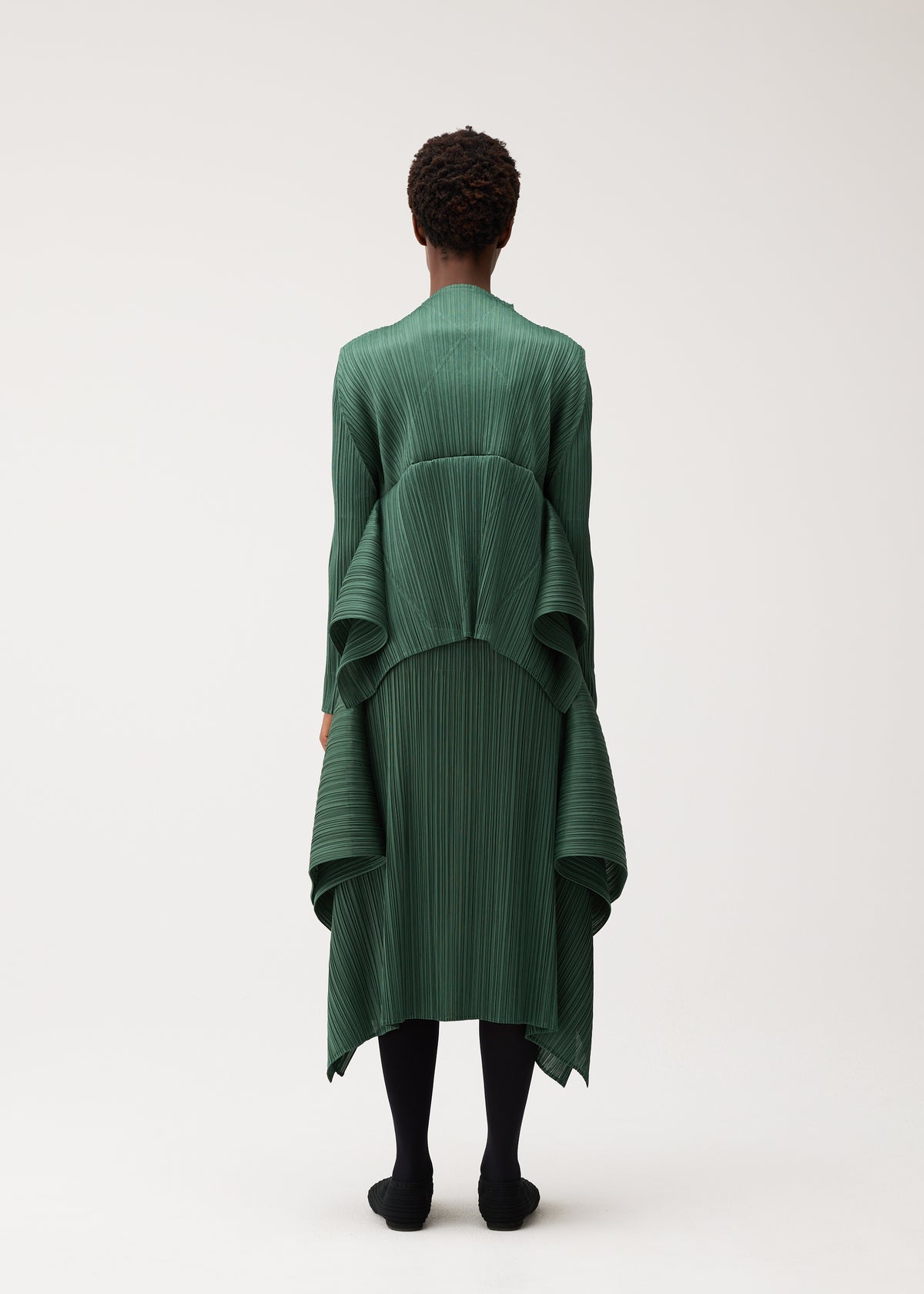 Zoomed view A person stands with their back to the camera, wearing the FLUTTER dress by PLEATS PLEASE ISSEY MIYAKE—a long, pleated dark green piece featuring draped, layered fabric. The outfit is paired with a distinctive cardigan for added texture and interest, along with black tights and shoes. The background is plain and light-colored.
