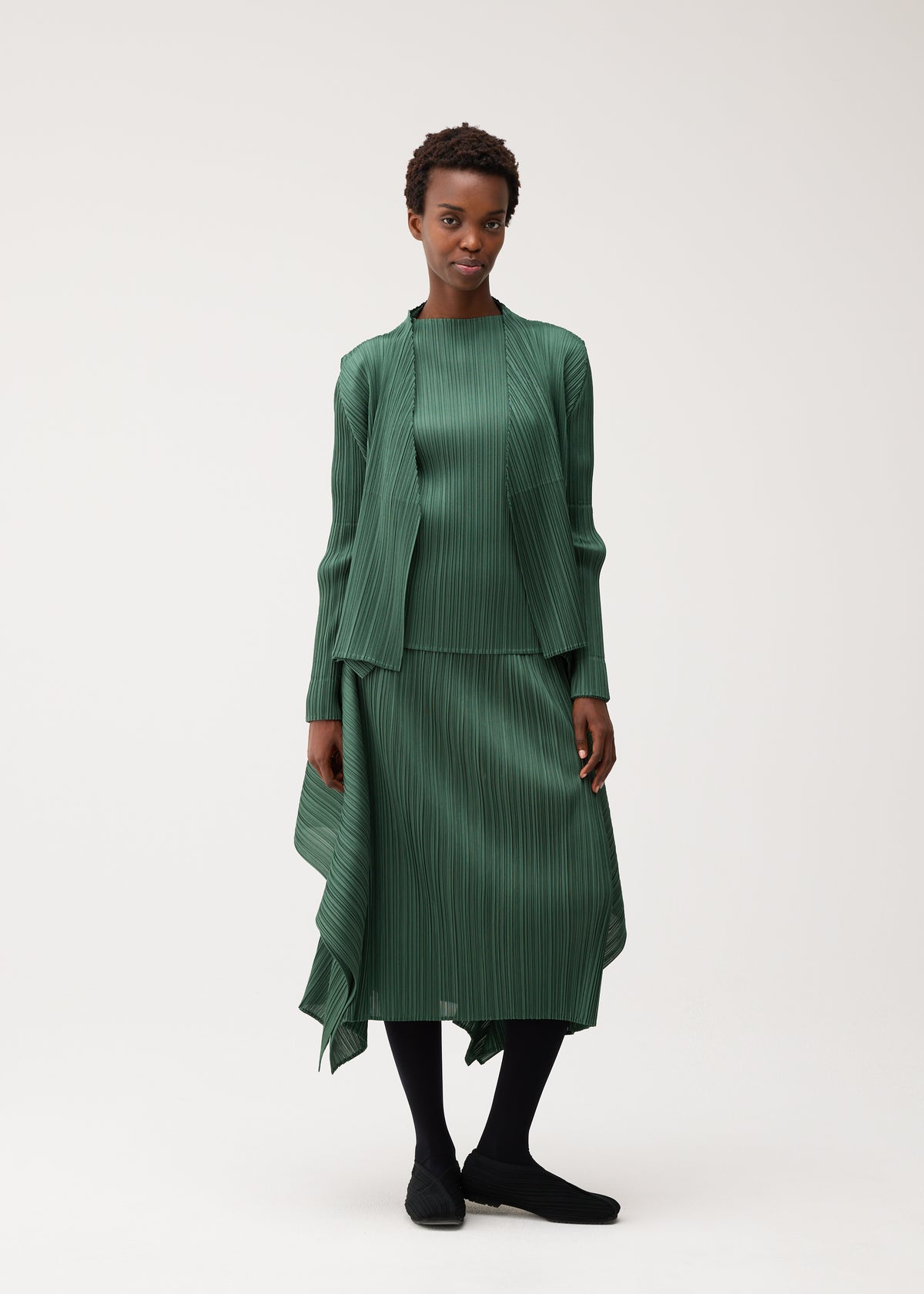 A person stands against a plain background wearing the FLUTTER matching pleated green top, jacket, and skirt from PLEATS PLEASE ISSEY MIYAKE, paired with black tights and black shoes. The look is completed with a long-sleeved cardigan that adds an elegant layer to the ensemble. view 9 of 9