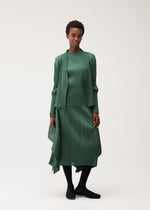 A person stands against a plain background wearing the FLUTTER matching pleated green top, jacket, and skirt from PLEATS PLEASE ISSEY MIYAKE, paired with black tights and black shoes. The look is completed with a long-sleeved cardigan that adds an elegant layer to the ensemble.