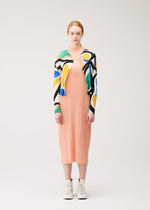 A woman faces forward against a light background wearing the ROLL peach pleated midi dress by PLEATS PLEASE ISSEY MIYAKE, a vibrant printed cardigan, and white lace-up ankle boots.
