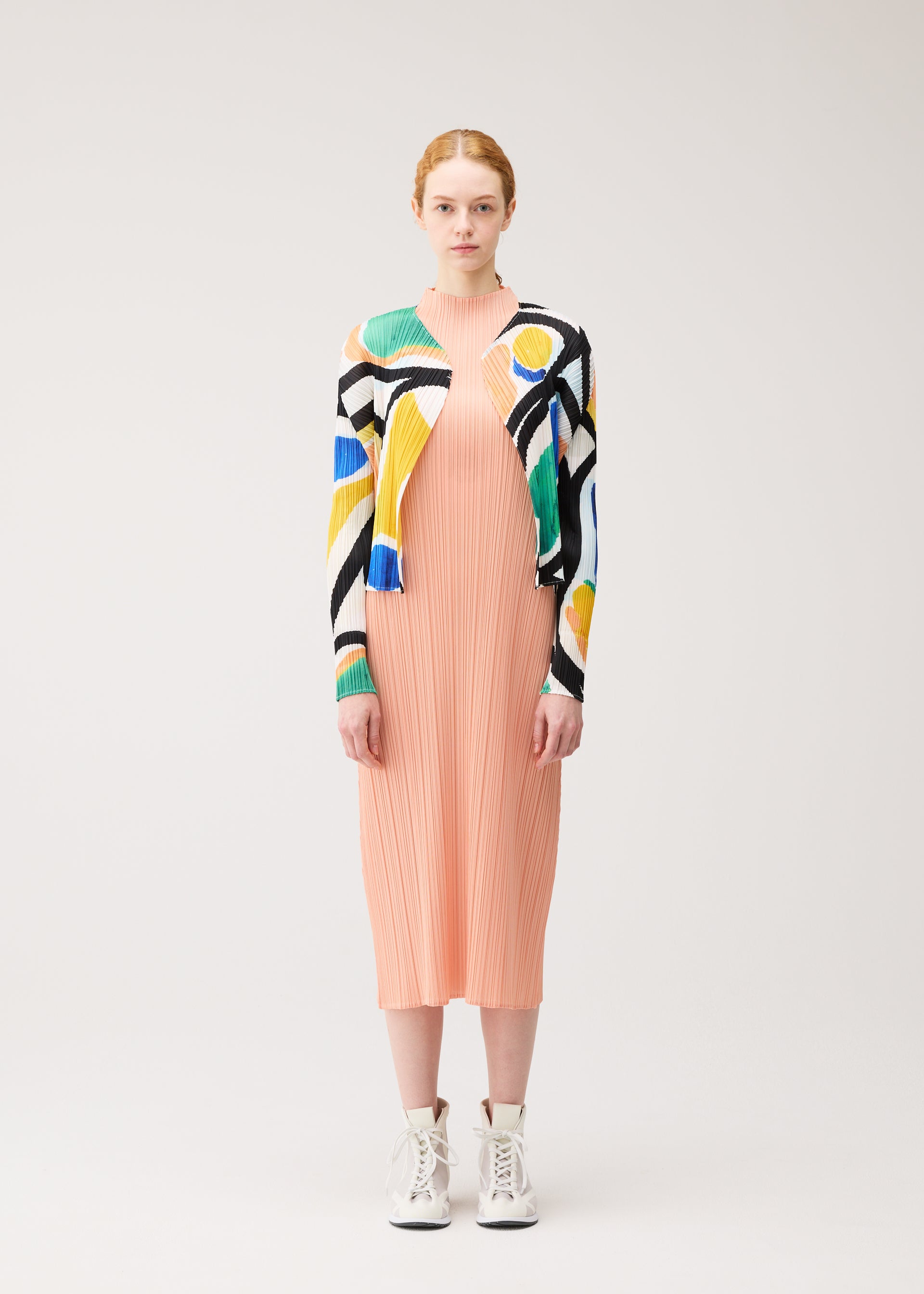 Zoomed view A woman faces forward against a light background wearing the ROLL peach pleated midi dress by PLEATS PLEASE ISSEY MIYAKE, a vibrant printed cardigan, and white lace-up ankle boots.