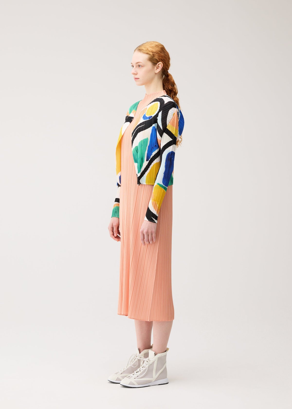 A woman with red hair in a ponytail stands sideways, wearing the ROLL peach pleated midi dress by PLEATS PLEASE ISSEY MIYAKE, a colorful printed cardigan, and white high-top sneakers against a plain light background. view 5 of 7
