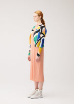 A woman with red hair in a ponytail stands sideways, wearing the ROLL peach pleated midi dress by PLEATS PLEASE ISSEY MIYAKE, a colorful printed cardigan, and white high-top sneakers against a plain light background.