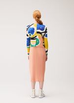 A person with long red hair in a braid stands facing away, wearing the PLEATS PLEASE ISSEY MIYAKE ROLL cardigan, a peach pleated midi skirt, and white high-top shoes against a plain light background.