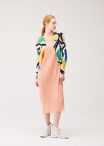 A woman with red hair in a low ponytail wears the ROLL peach pleated midi dress by PLEATS PLEASE ISSEY MIYAKE, a colorful printed cardigan, and white high-top sneakers. She stands slightly turned against a plain light background.