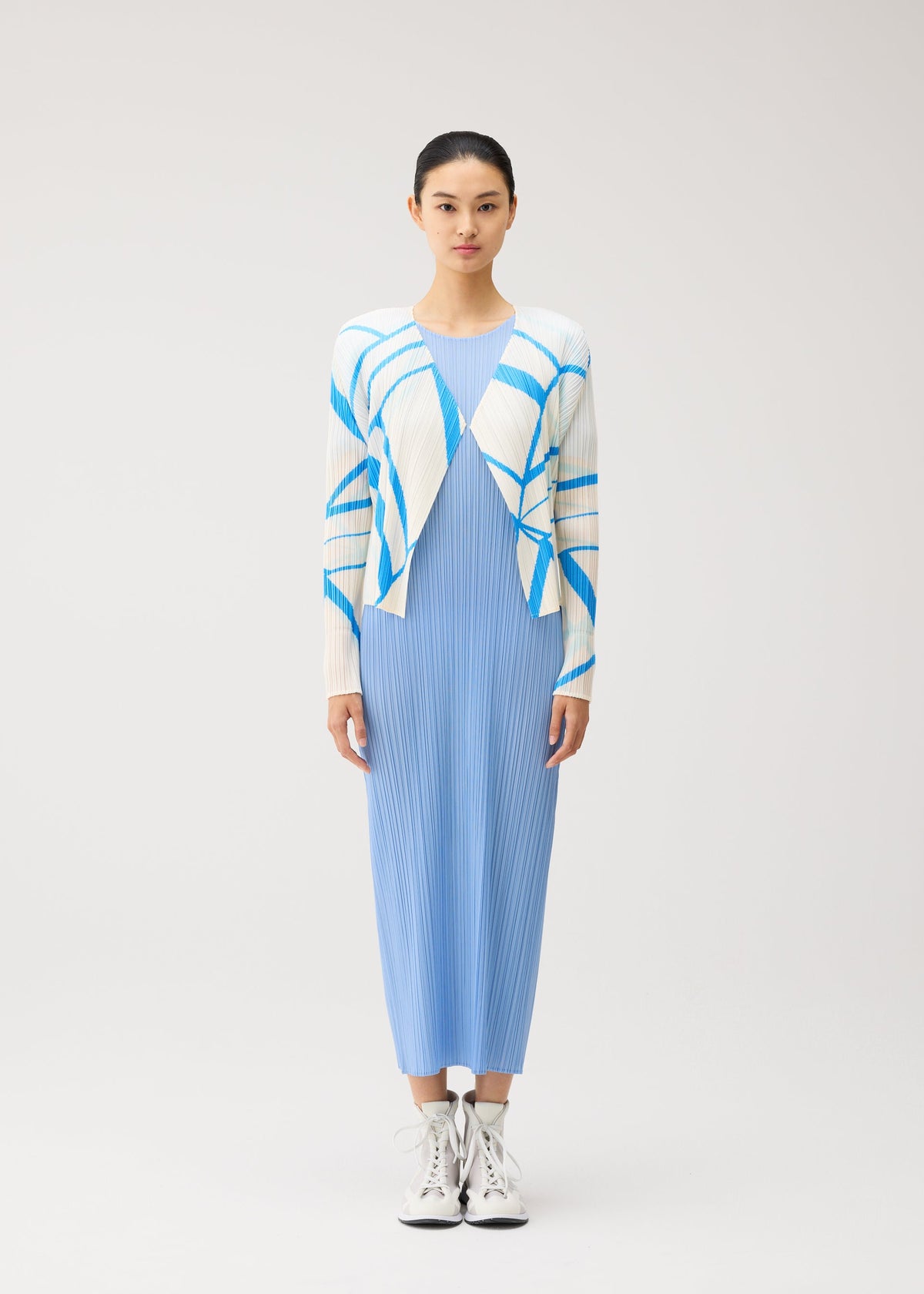 A woman stands facing forward, wearing the FOLD LINE long light blue pleated dress by PLEATS PLEASE ISSEY MIYAKE, styled with a minimalist white cardigan featuring blue stripes and paired with white lace-up boots, against a plain light background. view 4 of 7