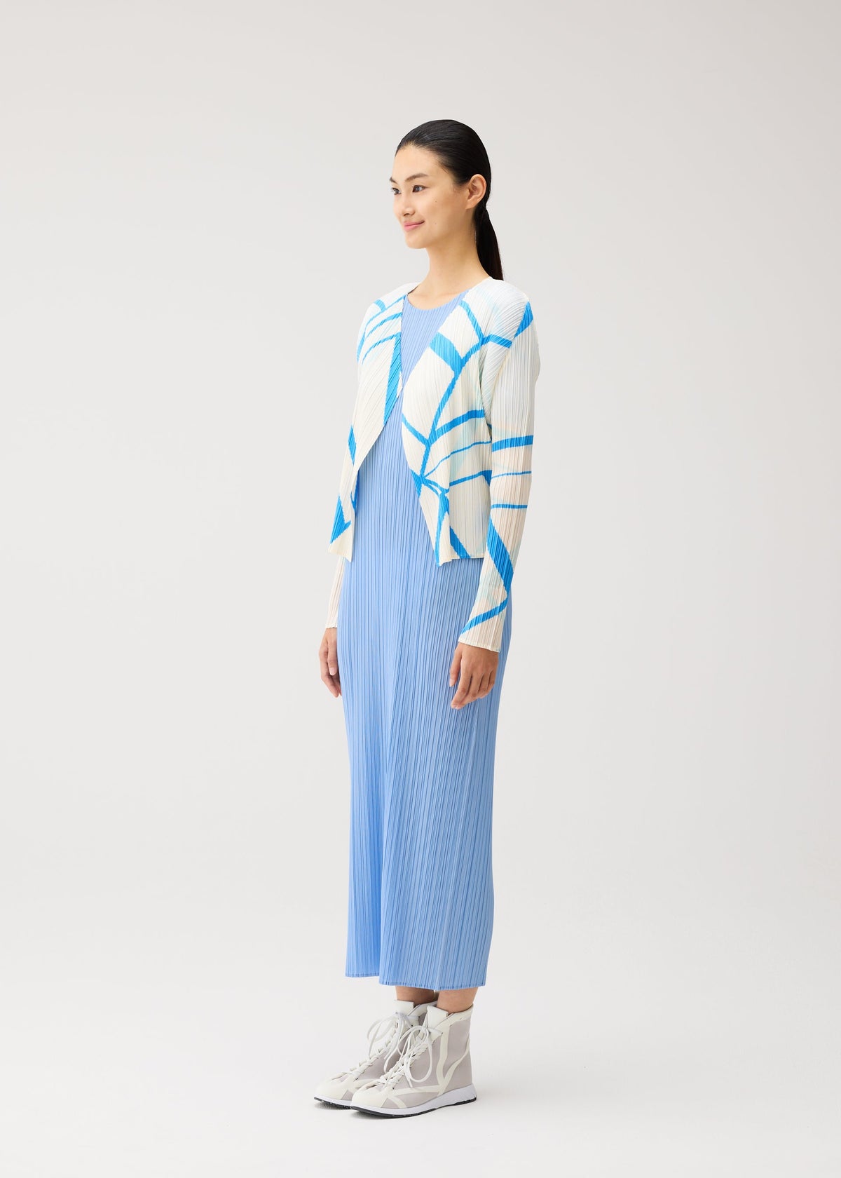 A woman stands in the FOLD LINE dress by PLEATS PLEASE ISSEY MIYAKE, showcasing its minimalist pleated light blue design, styled with a white and blue patterned cardigan and white high-top sneakers against a plain light background. view 5 of 7