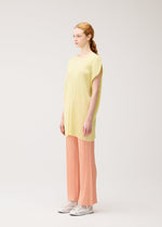 A woman with light skin and reddish hair stands in profile, wearing MONTHLY COLORS : JULY by PLEATS PLEASE ISSEY MIYAKE—a loose yellow pleated tunic, peach pleated wide-leg pants, and white sneakers—against a plain light background.