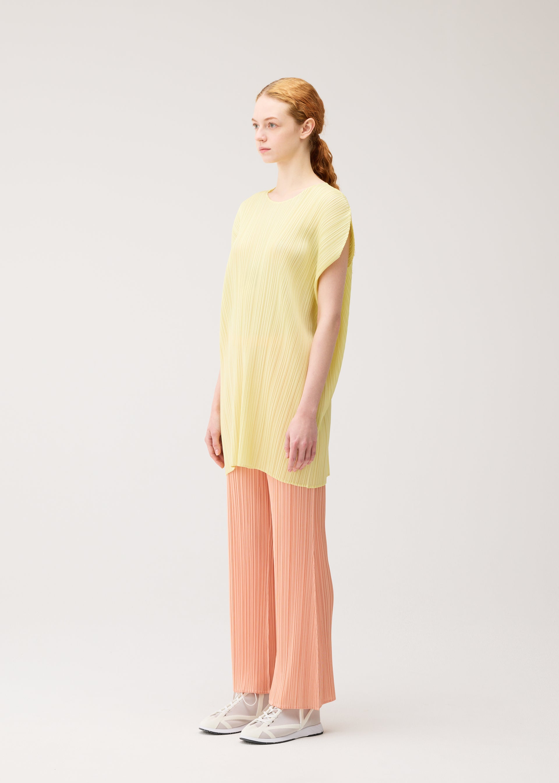 Zoomed view A woman with light skin and reddish hair stands in profile, wearing MONTHLY COLORS : JULY by PLEATS PLEASE ISSEY MIYAKE—a loose yellow pleated tunic, peach pleated wide-leg pants, and white sneakers—against a plain light background.