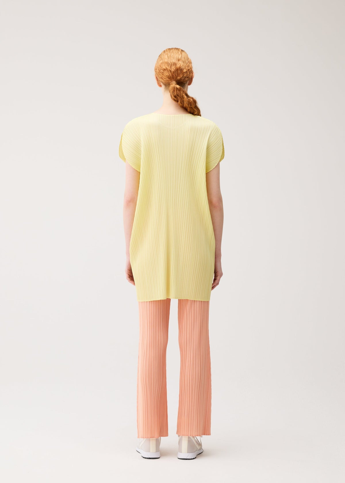 A person with red hair in a low ponytail is seen from behind, wearing the MONTHLY COLORS : JULY tunic and light peach pleated pants by PLEATS PLEASE ISSEY MIYAKE, paired with white shoes against a plain light background. view 10 of 11