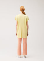 A person with red hair in a low ponytail is seen from behind, wearing the MONTHLY COLORS : JULY tunic and light peach pleated pants by PLEATS PLEASE ISSEY MIYAKE, paired with white shoes against a plain light background.