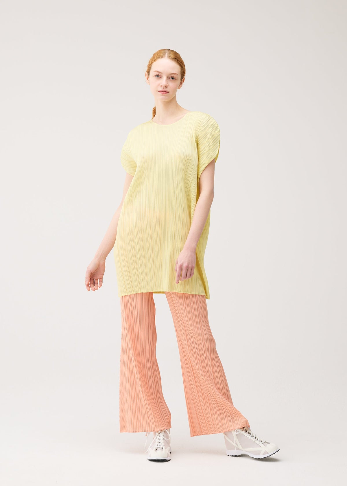 A model with light skin and long red hair wears PLEATS PLEASE ISSEY MIYAKE’s MONTHLY COLORS: JULY— a loose, yellow tunic with peach wide-legged pleated pants and white lace-up shoes, posed against a plain light background. view 11 of 11