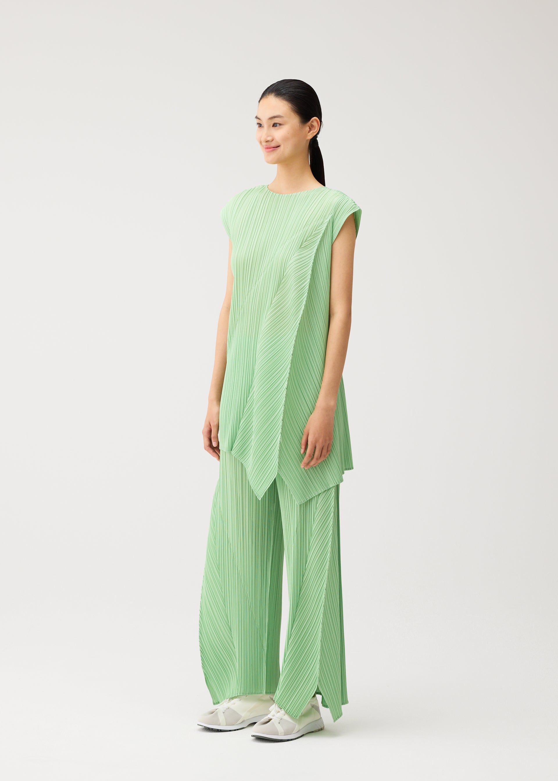 Zoomed view A person stands in a studio wearing the FOLD pleated round-neck sleeveless tunic and matching wide-leg pants in light green from PLEATS PLEASE ISSEY MIYAKE, paired with white shoes. The background is plain and light-colored.