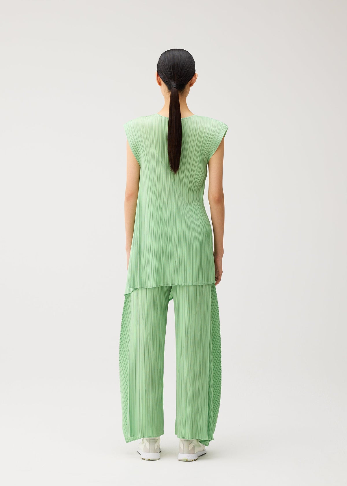 A person with long dark hair in a ponytail models the PLEATS PLEASE ISSEY MIYAKE FOLD set—a loose, pleated light green round-neck sleeveless tunic with matching wide-leg pants—standing facing away against a plain white background. view 7 of 8