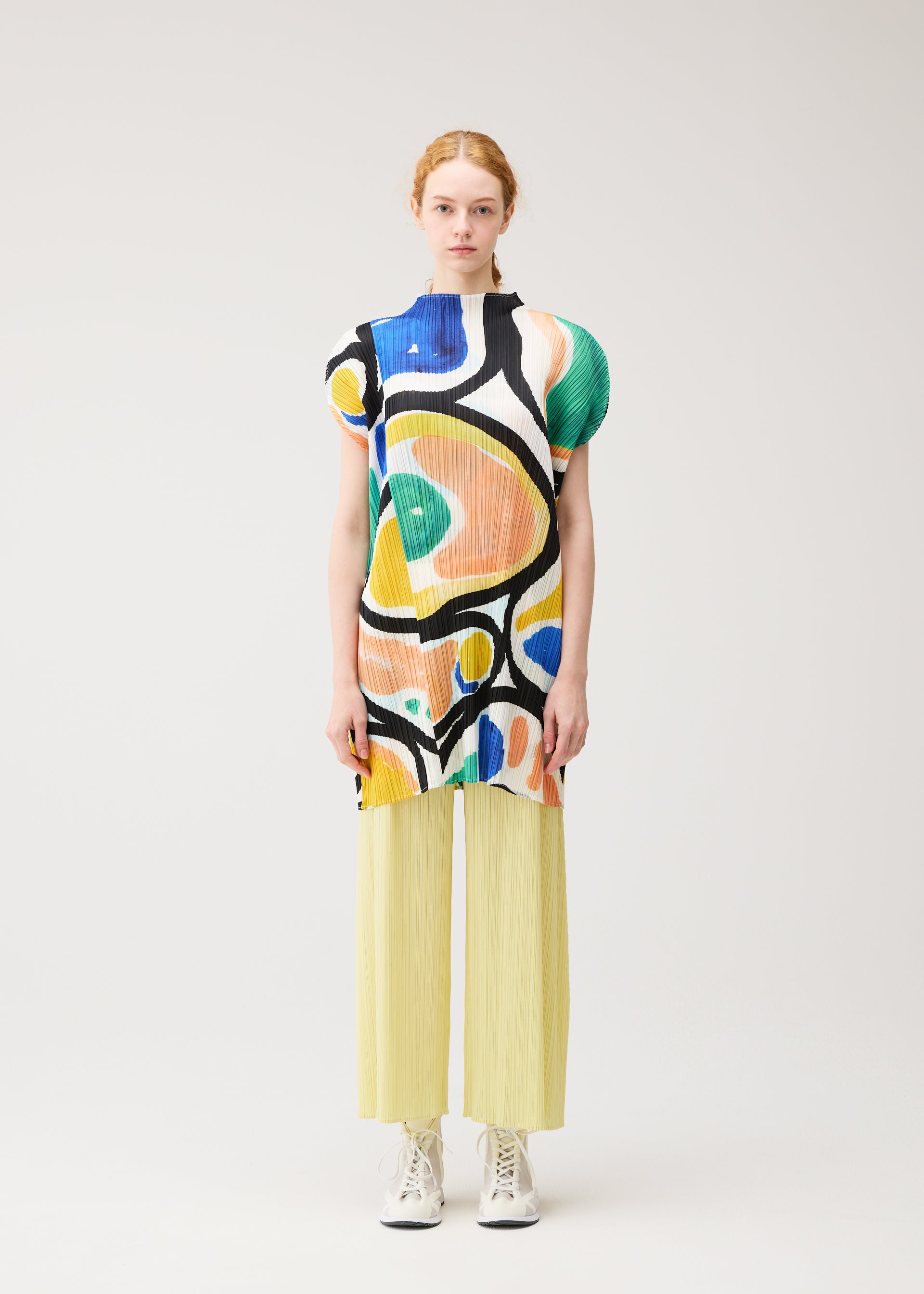 Zoomed view A woman stands against a plain background wearing the ROLL tunic by PLEATS PLEASE ISSEY MIYAKE, featuring a sushi roll print, paired with yellow pleated pants and white sneakers. She faces forward with a neutral expression.
