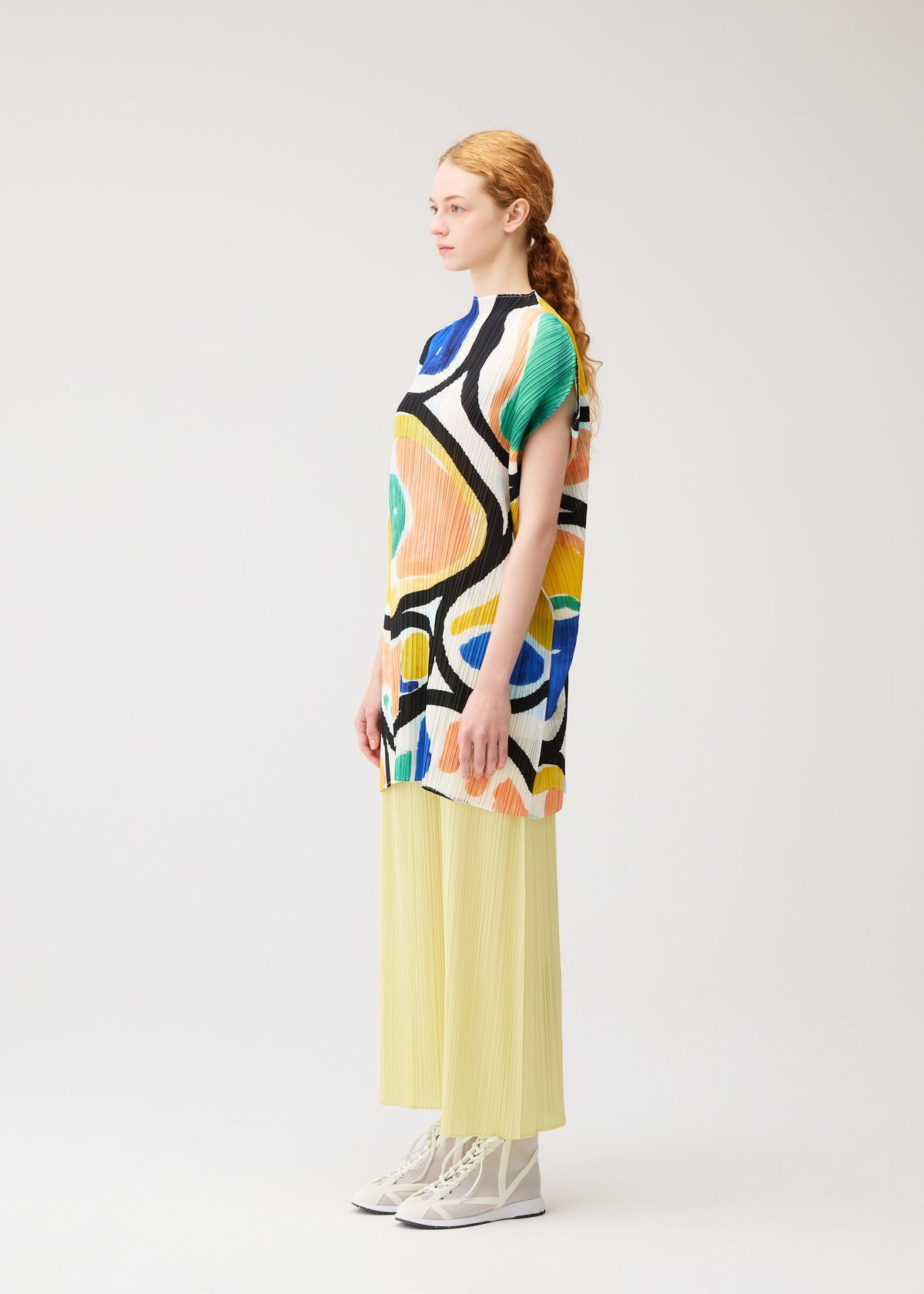 A woman with red hair in a low ponytail stands sideways, wearing the ROLL tunic by PLEATS PLEASE ISSEY MIYAKE, pale yellow pleated wide-leg pants, and white high-top sneakers against a plain light background. view 6 of 8