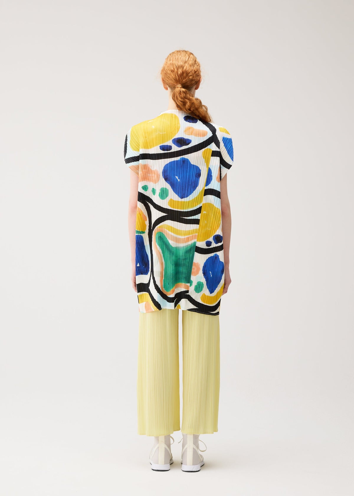 A person with long red hair in a braid wears the ROLL tunic by PLEATS PLEASE ISSEY MIYAKE, paired with light yellow pleated pants and white shoes, standing facing away against a plain light background. view 7 of 8