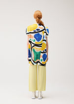 A person with long red hair in a braid wears the ROLL tunic by PLEATS PLEASE ISSEY MIYAKE, paired with light yellow pleated pants and white shoes, standing facing away against a plain light background.